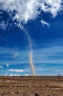 A narrow tornado crosses a dry plain, the sky is clear with scattered clouds and the mountains are