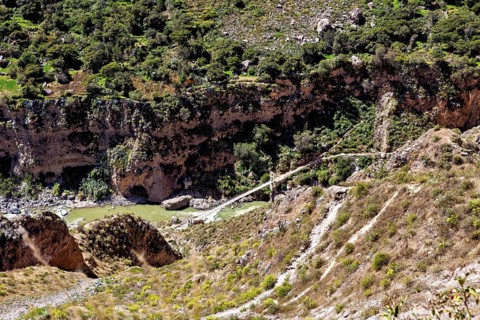 Wooden bridge across river in an overgrown valley surrounded by rocks and thick greenery, The