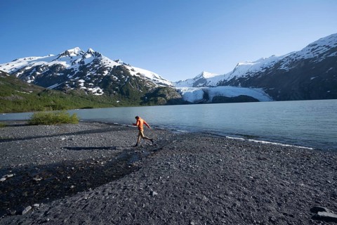 Young man jumping on a pebble beach on a glacial lake, Snowy Mountains and Portage Glacier on