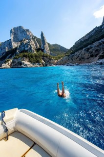 Young woman jumping from a boat into the water, picturesque rocky coast, cliffs with L'Aguglia rock
