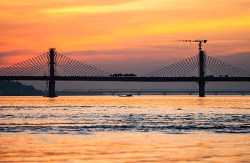 A boat sails on the river Brahmaputra during sunset, with the silhouette of a cable-stayed bridge