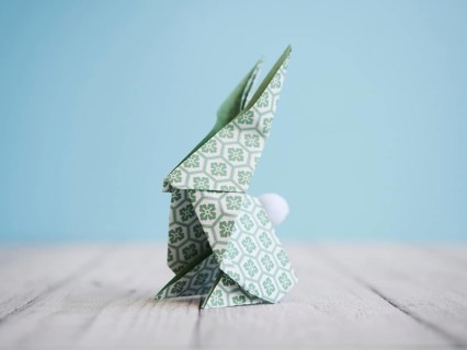 Green origami bunny standing on a white wooden table with a light blue background, representing