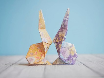 Two colorful origami bunnies are facing each other, touching noses, on a wooden surface with a