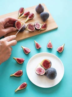 Whole and sliced fresh figs on a blue background, with hands cutting them on a wooden board