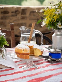 Slice of lemon cake with powdered sugar on a glass stand, pitcher of lemonade, flowers, and