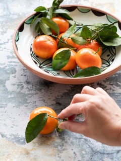 Fresh tangerines with leaves are being picked up from a ceramic bowl on a textured surface