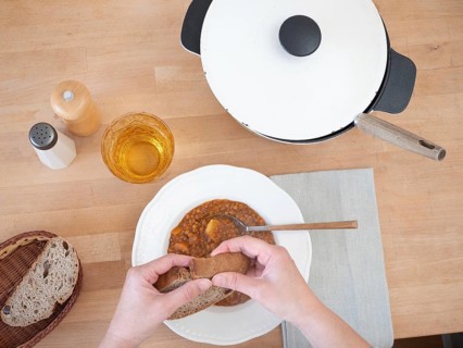 Hands breaking slice of bread over bowl of lentil soup, served with drink, salt and pepper, and