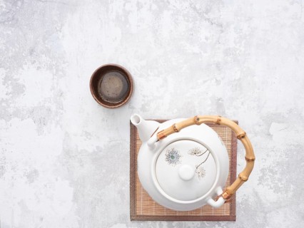 White teapot with bamboo handle and small wooden teacup on bamboo mat, ready for traditional asian