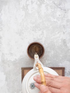 Hand gracefully pours tea from a white ceramic teapot with bamboo handle into a small brown bowl on
