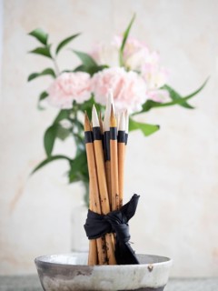 Calligraphy brushes are tied with a black ribbon and placed in a ceramic bowl, with pink carnations