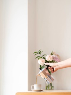 Hand pouring coffee from moka pot into cup on wooden table, with flowers in background, in