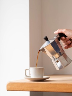 Hand pouring coffee from a moka pot into a cup, placed on a wooden table, creating a cozy and