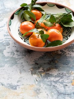 Vibrant tangerines with green leaves arranged in a decorative ceramic bowl, set against a rustic