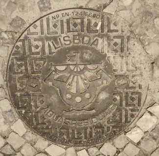 Engraved manhole cover on cobblestones with decorative patterns and a city coat of arms, Lisbon,