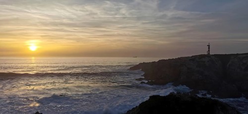 A lighthouse stands on cliffs in front of a sunset over the sea, hiking the Fisherman's Trail, Rota