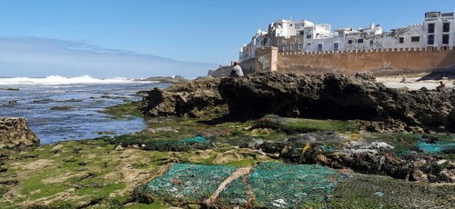 Rocky coast with city walls and view of Essaouira blue sky and algae in daylight, a man sitting on