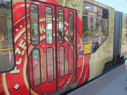 A tram with Pilsner Urquell painting stops at a sunny city stop, Pilsen, Czech Republic
