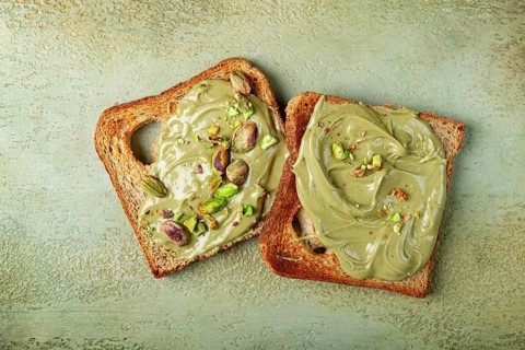Two slices of toasted bread, topped with pistachio paste and sprinkled with pistachios, spread on a