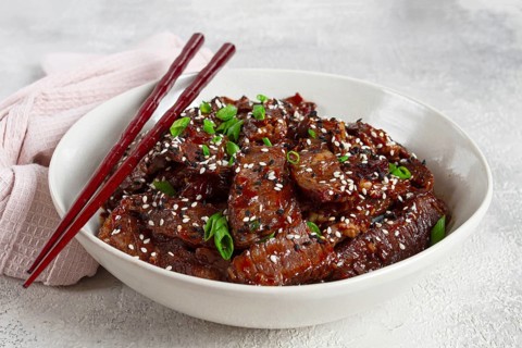 Crispy beef, with green onions and sesame seeds, in a bowl with chopsticks, on a textured surface,