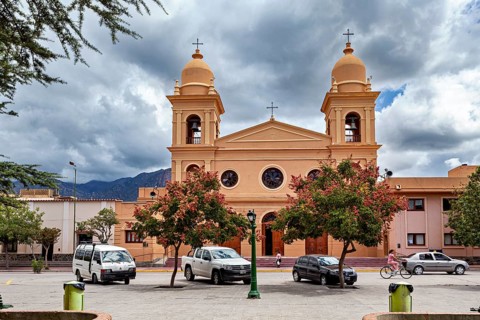 Church façade with two bell towers surrounded by trees and cars, under a dramatic sky, the Catedral