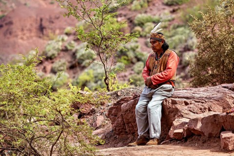 A traditionally dressed man sits thoughtfully on a rock in a desert landscape, a local in the rock