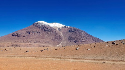 A bizarre mountain with a snow-capped summit rises above the dry desert, the landscape and colorful