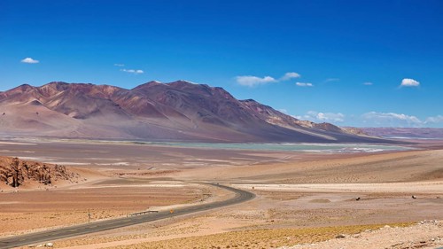 A long road snakes through an arid desert landscape with distant mountains, the countryside and