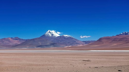 A snow-covered volcano rises above a dry, vast desert under clear skies, the landscape and colorful