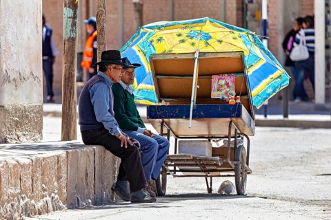 Two men resting under a blue parasol next to a cart, people in the streets of San Antonio de los