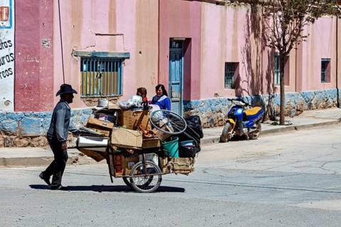 Man pulls carts full of packages past a colorful building, people in the streets of San Antonio de