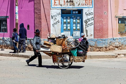 Man wearing hat pulls a full cart past a pink building, people in the streets of San Antonio de los