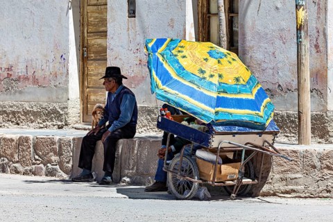 Man and child sitting under a blue parasol next to a cart, people in the streets of San Antonio de