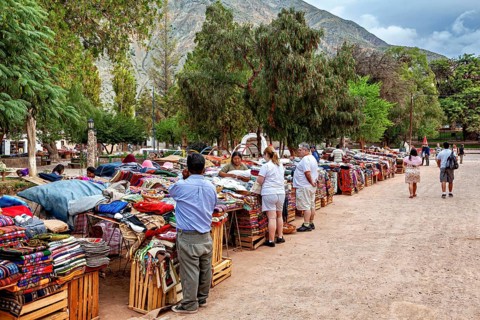 People check out textile stalls at a lively market against a mountain backdrop, tourist and