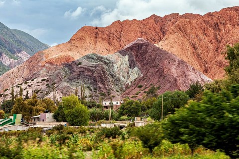 Colourful mountains rise from a green, overgrown desert under cloudy skies, the landscape and