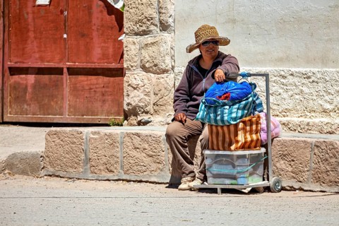 Man wearing hat sitting on the side of a road next to a loaded cart, people in the streets of San