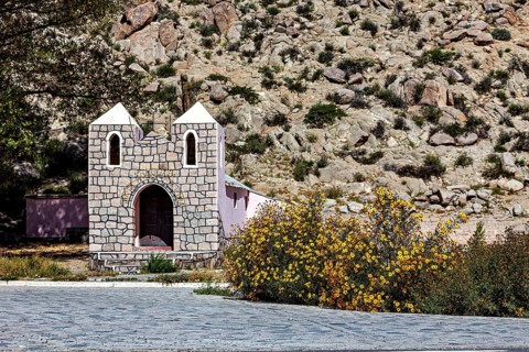 Small stone house in a desert area with yellow flowers in the foreground against a rocky mountain