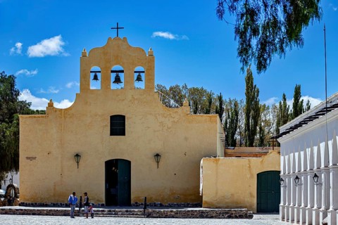 Yellow church with bell tower under blue sky, people on stairs surrounded by trees, Iglesia San