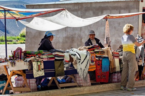 Two woman at market stalls surrounded by handmade blankets, interacting with a customer, tourist