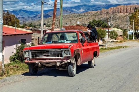 Red, slightly damaged pickup truck on a country road with a man and mountain scenery, old cars in