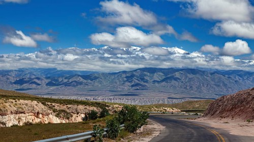 Curvy road leads through a vast landscape with mountains and clouds in the sky, roads through the