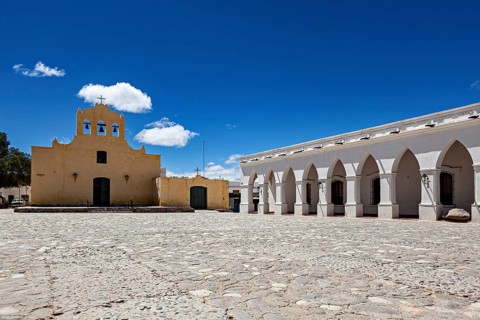 Square with yellow church and white arcades under bright blue sky, Iglesia San José de Cachi in