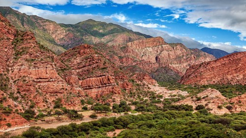 An impressive mountain landscape with red rocks, lush vegetation and a partly cloudy sky, the