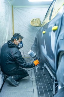 Auto body technician wearing mask and gloves sprays primer onto a car door panel inside a