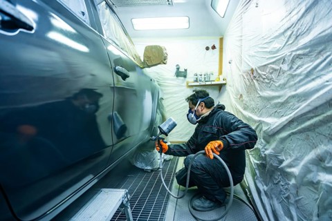 Mechanic wearing respirator mask and orange gloves spraying dark car in a professional paint booth,