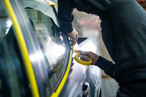 Auto body technician hands applying yellow masking tape around car window trim in a workshop,