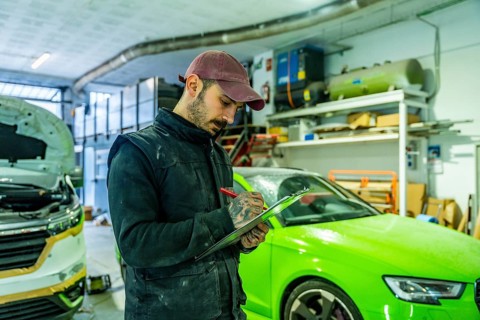 Auto body technician wearing a cap and overalls writing on a checklist, inspecting vehicles in a
