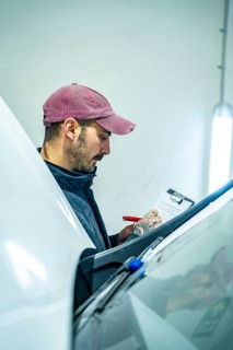 Auto technician wearing a cap writing details on a clipboard while inspecting vehicle condition in