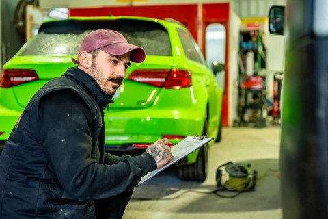 Automotive technician crouching and writing notes on a clipboard during a vehicle inspection or