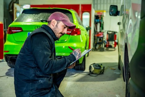 Auto body technician wearing a cap and overalls making notes on a clipboard while inspecting a