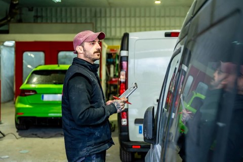 Mechanic holding a clipboard and pen while inspecting a commercial van in a car body and paint